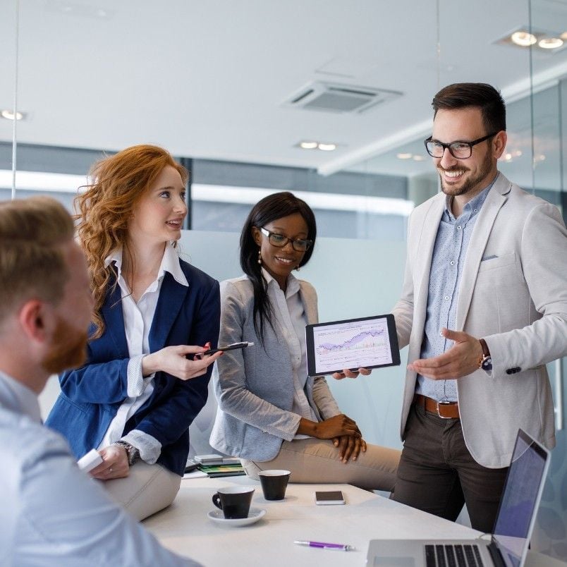 A group of four professionals in a modern office discuss a Stakeholder Map displayed on a tablet held by a smiling man in glasses, while others listen and engage around a table with coffee cups and documents.