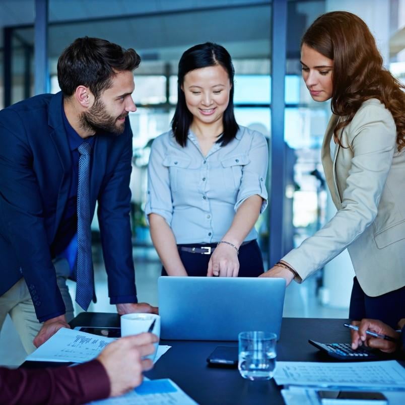 Three business professionals stand around a table in a modern office, looking at a laptop screen and discussing work. Papers, a coffee cup, and water glasses fill the table as they review Epic Examples of success together.