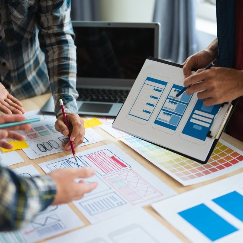 People collaborate at a desk covered with website wireframe sketches and color swatches, using Project Management Software for Small Businesses to organize ideas. One person holds a clipboard while others discuss layouts in an efficient planning meeting.