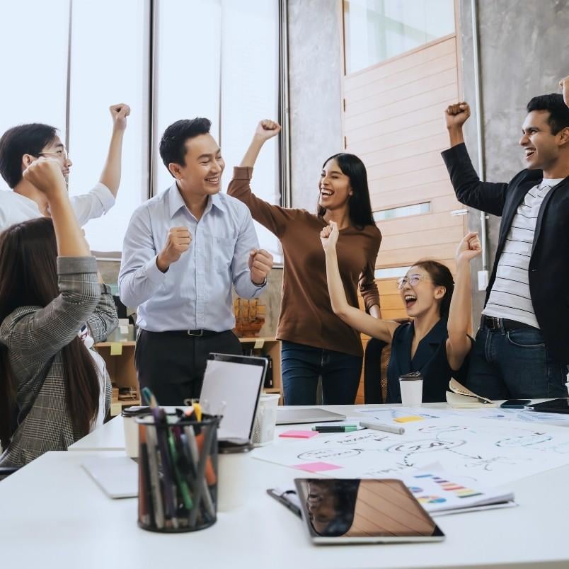 A group of six coworkers cheer and celebrate together around a desk covered with papers, laptops, and office supplies in a bright, modern office space—creating one of those epic examples of teamwork and success.