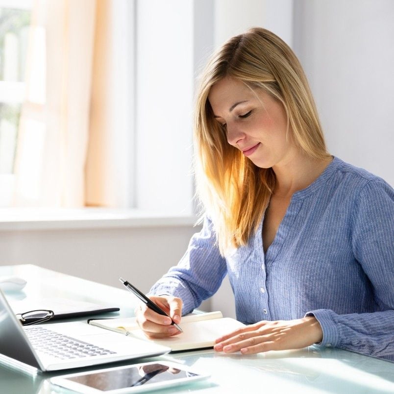 A woman with blonde hair in a blue shirt sits at a desk, writing in a notebook on how to write an epic. A laptop, phone, and pen are on the desk as sunlight streams through a window in the background.