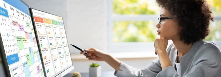 A woman sits at a desk, looking at two large computer monitors displaying colorful digital calendars and schedules on Project Management Software for Small Businesses. She holds a pen and appears focused while working in a bright office.