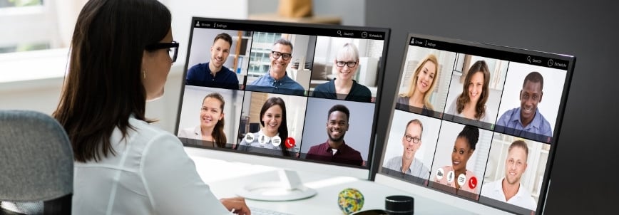 A woman sits at a desk participating in a video conference on two computer monitors, each displaying several people in a virtual meeting grid—a scene that highlights project management for remote teams.