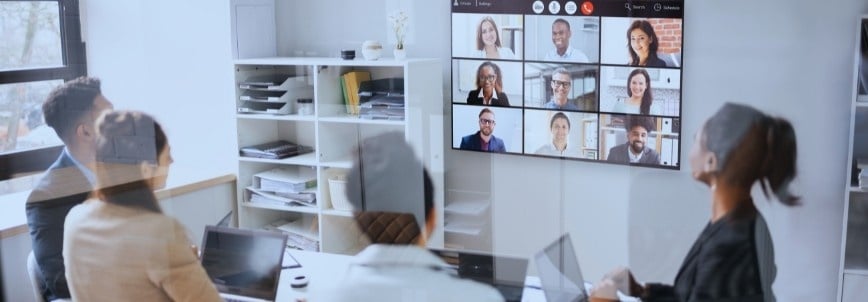 Four people in a conference room engage in virtual collaboration, facing a large screen with several participants on a video call. Laptops and office supplies are spread across the table.