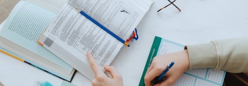 A person studies at a table with an open textbook, worksheet, and blue pen in hand, seeking epic examples. Glasses and a bottle of hand sanitizer are also visible on the white surface.