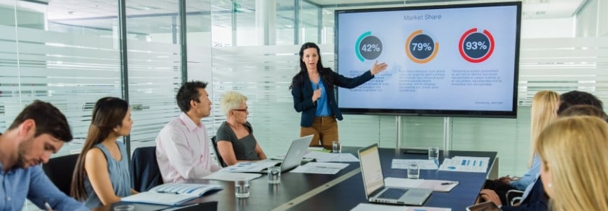 An Agile Program Manager stands and presents market share data on a large screen to a group of people seated around a conference table in a modern office meeting room.