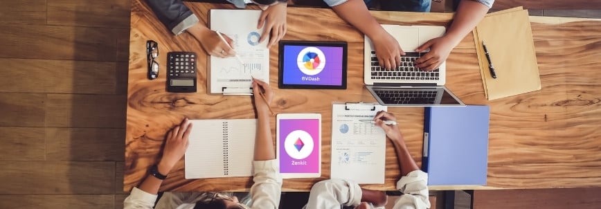 Four people collaborate at a wooden table with laptops, tablets, charts, and calculators, comparing data for business reports. Their discussion includes analyzing project management tools like BVDash vs. Zenkit from an overhead perspective.