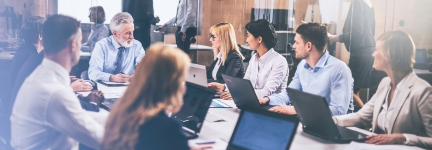 A diverse group of business professionals sit around a conference table with laptops, discussing strategies and reviewing an Epic Template in a modern office setting.