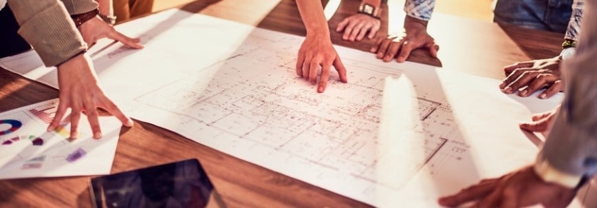Several people gather around a table, examining and pointing to a large architectural blueprint spread out before them, discussing details as part of a collaborative design meeting with an emphasis on project tracking.