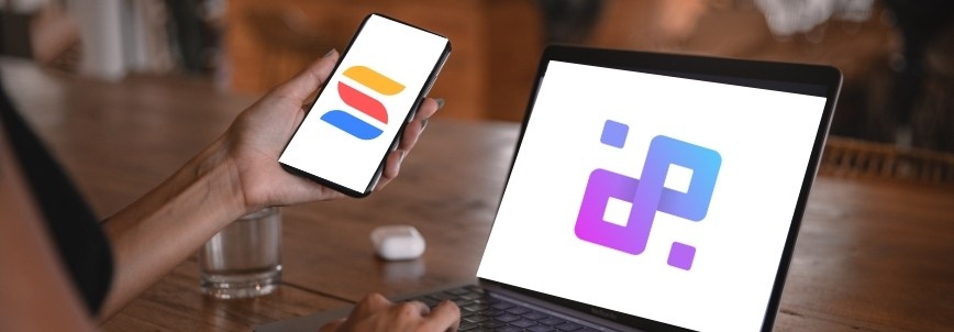 A person holds a smartphone displaying the Infinity vs. SmartSuite logo, while using a laptop that shows a vibrant purple and blue logo. A glass of water and wireless earbuds rest on the wooden table nearby.