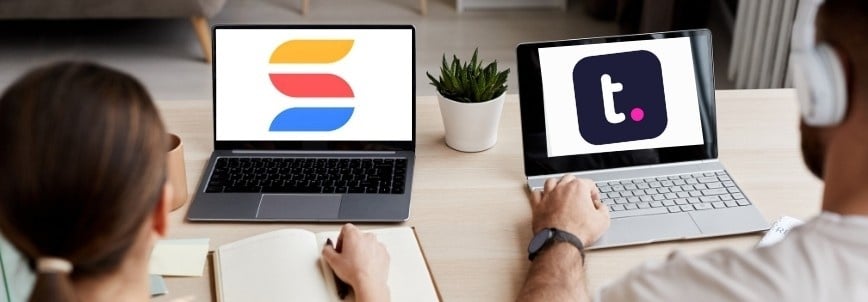 Two people sit at a table with open laptops—one displaying the SmartSuite logo, the other showing Teamwork.com's signature "t" and pink dot. A notebook and plant add to their workspace as they compare SmartSuite vs Teamwork.com.