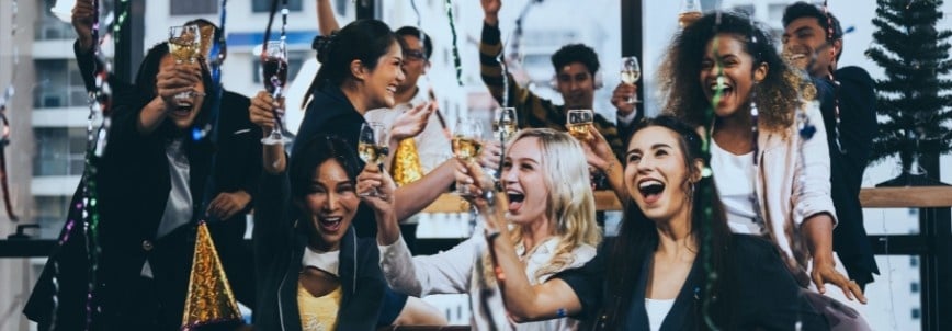 A group of people in festive attire cheer, laugh, and raise glasses at a lively indoor party, celebrating their successful project management together amid streamers and party hats in a joyful atmosphere.