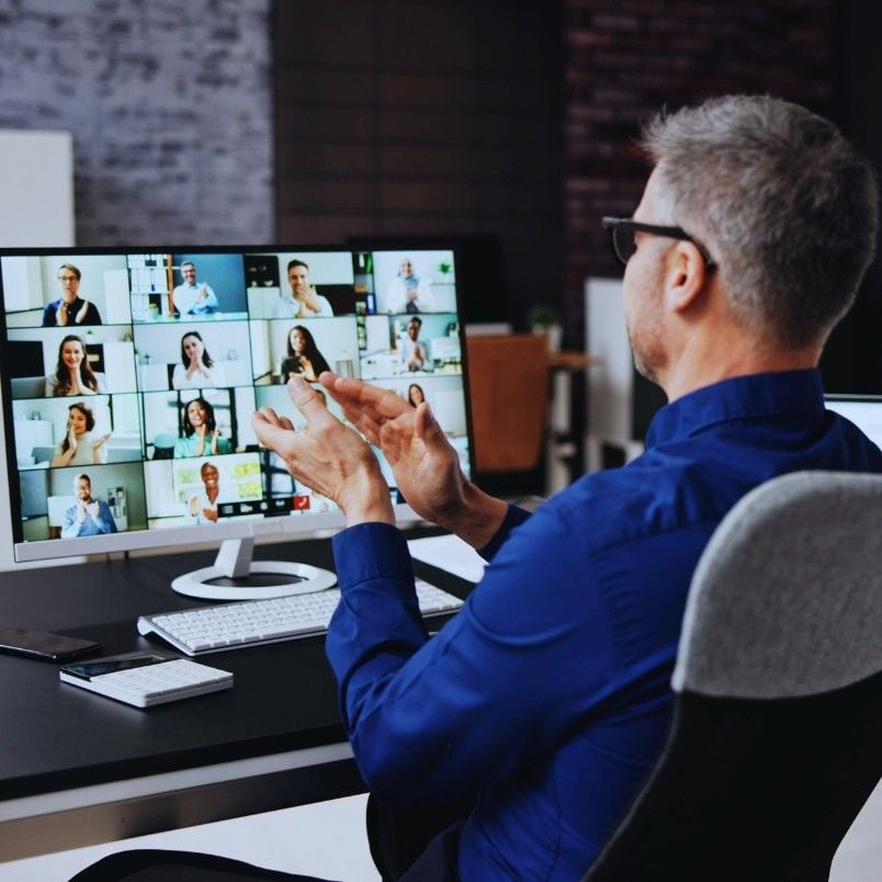 A man in a blue shirt sits at a desk, clapping while participating in a virtual collaboration with multiple people displayed on a large computer screen.