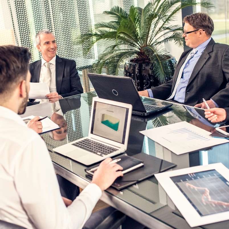 Four men in business attire sit around a modern glass conference table with laptops, graphs, and documents, discussing project management for remote teams. Large windows and a potted plant are visible in the background.