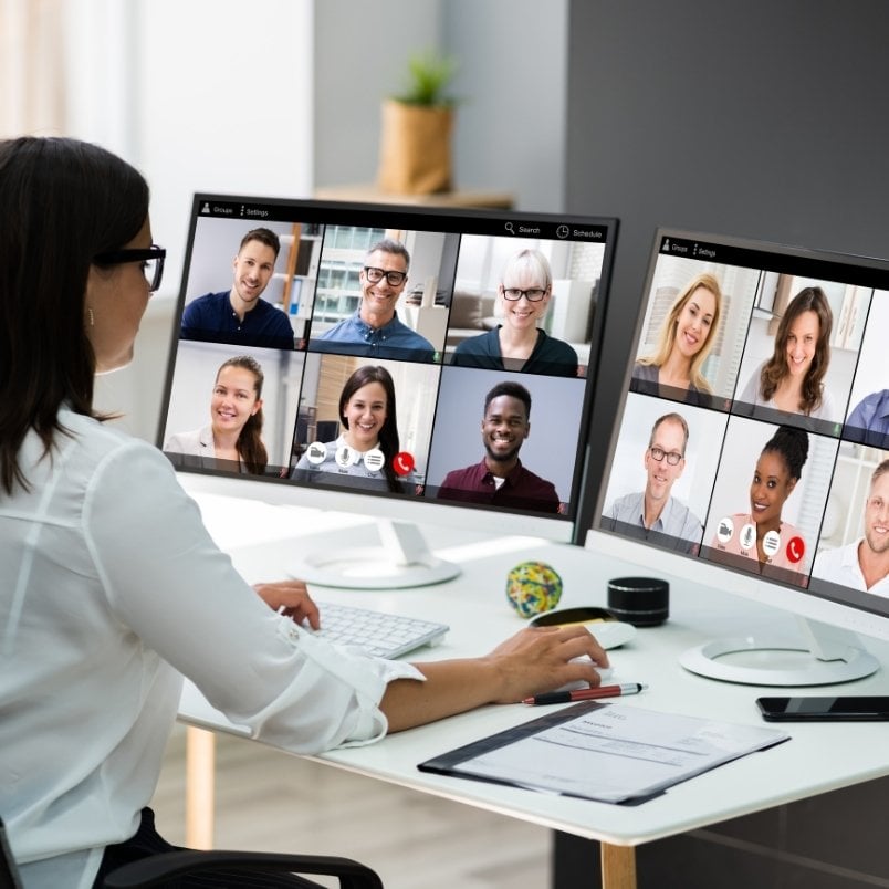 A woman sits at a desk using two monitors, each displaying a video call with several people on screen, engaged in project management for remote teams in a modern office setting.