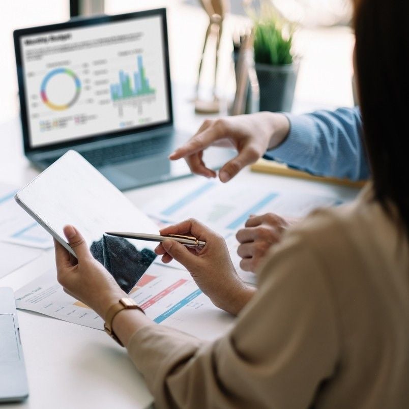 Two people review a tablet with charts and documents, while a laptop displaying colorful graphs, financial data, and a stakeholder map sits on the desk in a modern office setting.