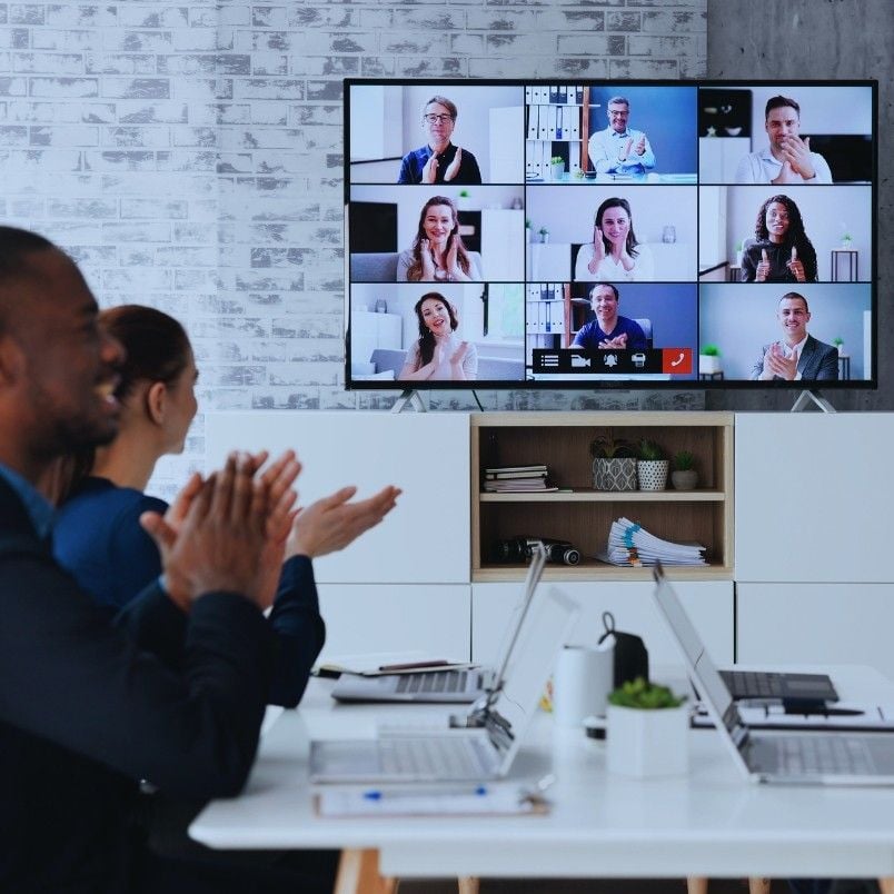 People sit at a table with laptops, clapping and smiling during a video conference. On a large screen, multiple people appear in a grid layout, also applauding, showcasing successful virtual collaboration.
