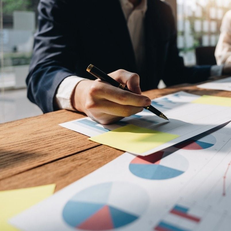 A person in business attire writes on a notepad at a desk with financial charts and graphs, including pie charts, bar graphs, and a stakeholder map, spread out on the table.