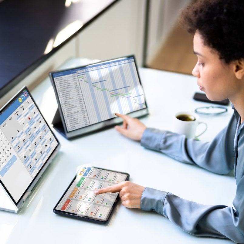 A person sits at a desk using a tablet and two monitors displaying a calendar, project management charts, schedules, and What Is An Epic In Scrum. A cup of coffee is nearby. The focused individual interacts with the digital screens.