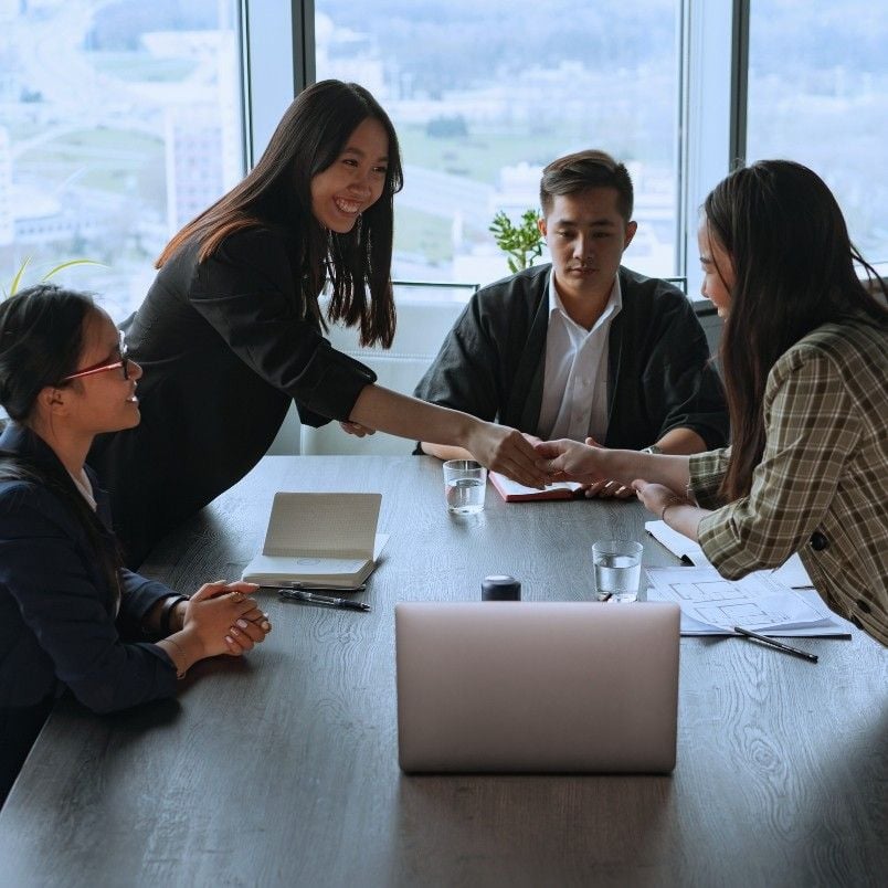 Four people sit around a conference table with laptops and documents; two women are shaking hands and smiling while the others watch, suggesting an Epic Example of a successful business meeting in a modern office with large windows.