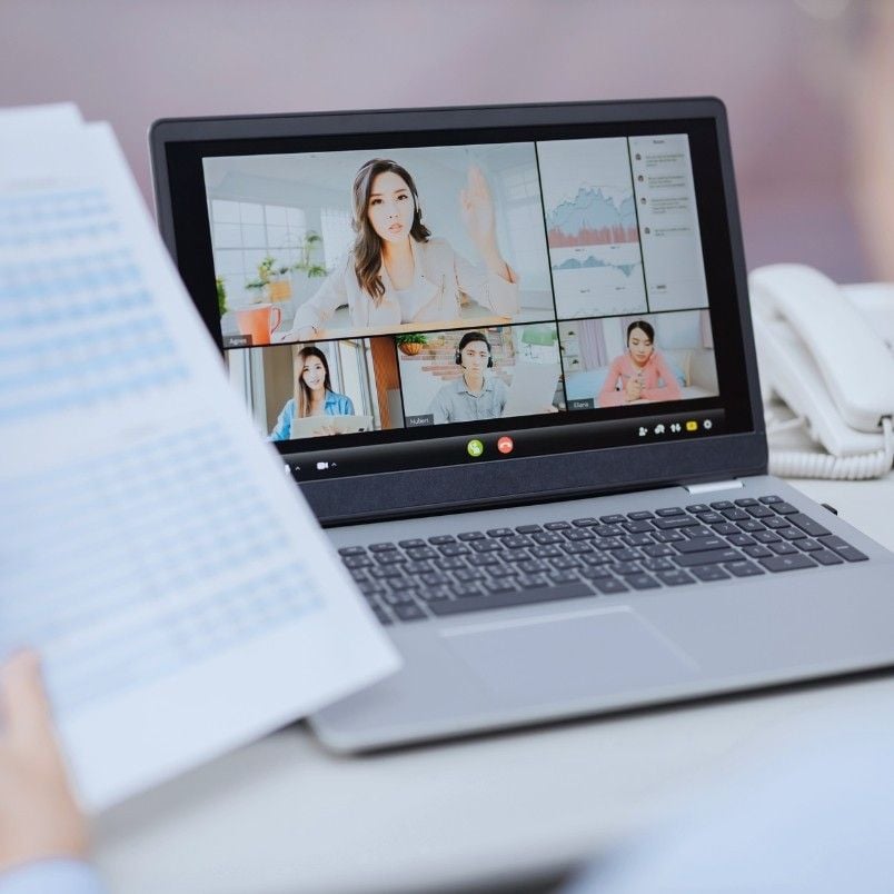 A person holds documents near a laptop displaying a video conference with four people in their own windows, highlighting virtual collaboration. Charts are visible on the screen, and a phone rests next to the laptop on the desk.