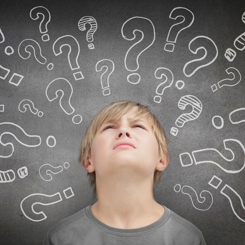 A young boy looks upward with a puzzled expression, standing in front of a chalkboard covered with large, white question marks—reflecting the confusion of choosing between smartsuite vs. zenkit.
