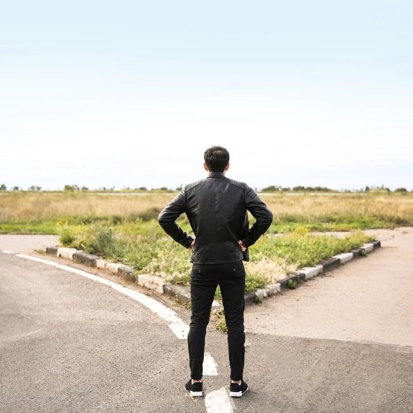 A man in a black jacket and pants stands at a fork in the road, as if pondering a choice like smartsuite vs. zenkit, gazing toward an open grassy field under a clear sky.