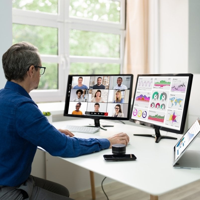 A person in a blue shirt sits at a desk with two monitors and a laptop. One screen shows a video conference for project management for remote teams; the other displays various colorful graphs and charts.
