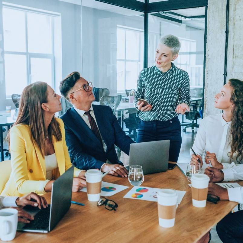 A group of four people sit at a conference table with laptops and charts, listening to a standing woman who is speaking and holding a phone—an epic example of teamwork during a meeting in a modern office.