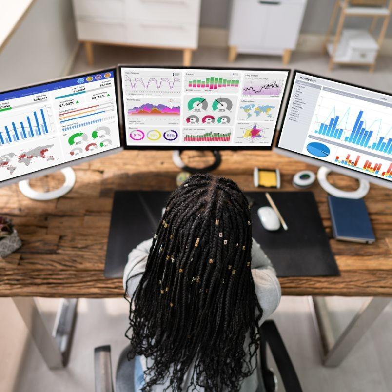 A person with braided hair sits at a desk using three computer monitors displaying colorful data charts, graphs, and Project Progress Tracking in BVDash in an office setting.