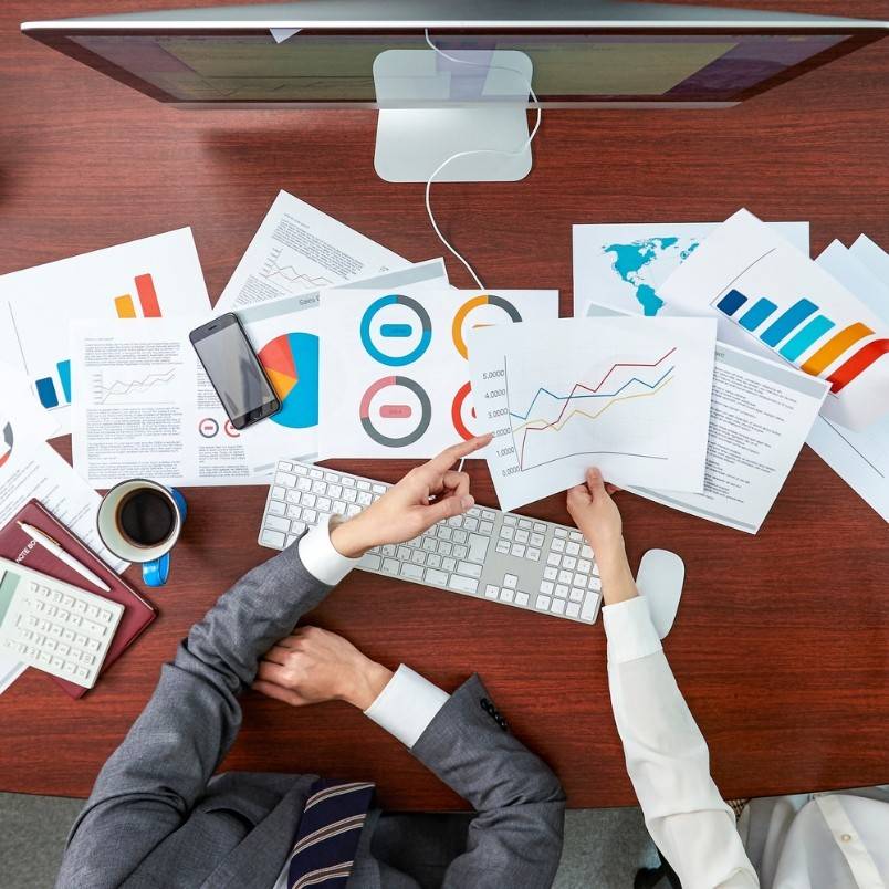 Top view of two people at a desk, analyzing charts and graphs for Project Progress Tracking in BVDash, with a computer, keyboard, phone, calculator, coffee cup, and office supplies spread out on the wooden surface.