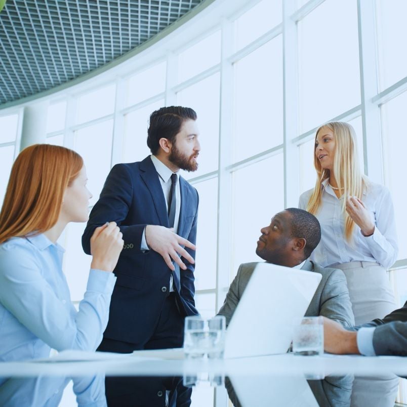 Four business professionals discuss SmartSuite vs. Hive around a table in a modern office with large windows; one man speaks as others listen attentively, with a laptop and glasses of water on the table.