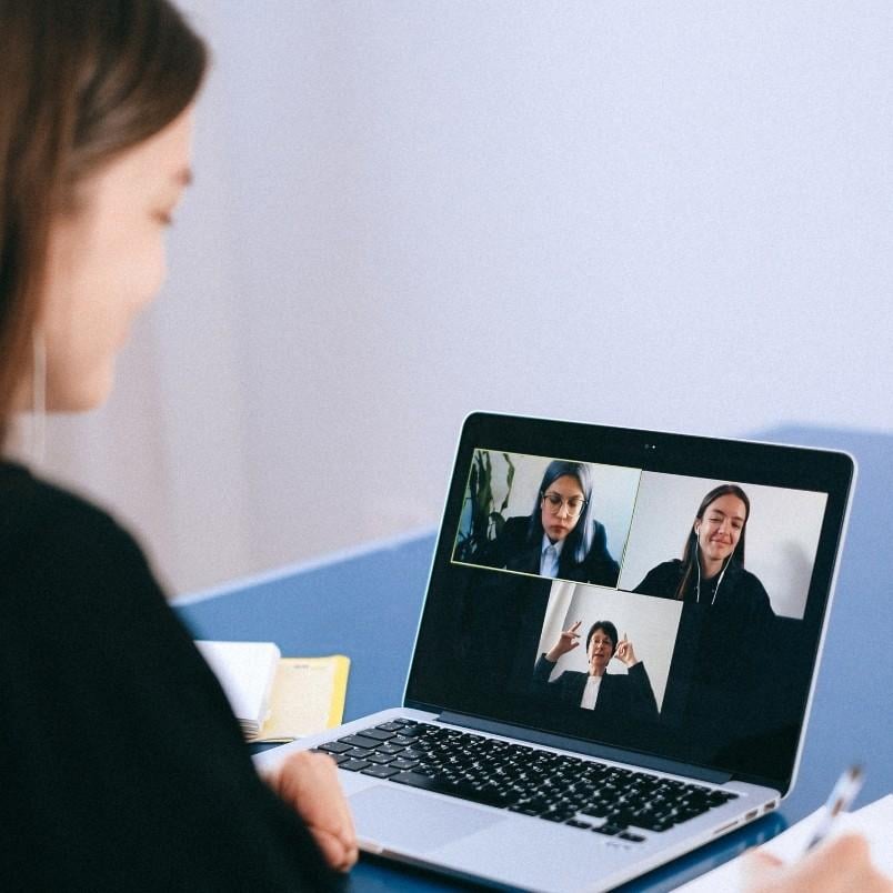 A person engages in virtual collaboration during a video call on a laptop with three people, each in their own window. A notebook and pen sit beside the laptop, ready for taking notes.
