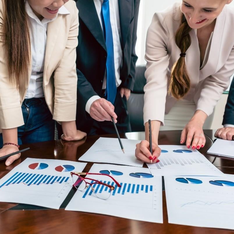 Four people in business attire review and discuss printed graphs, charts, and a Stakeholder Map on a table. Two women are smiling while pointing at the charts with pens. A pair of glasses rests on top of the papers.