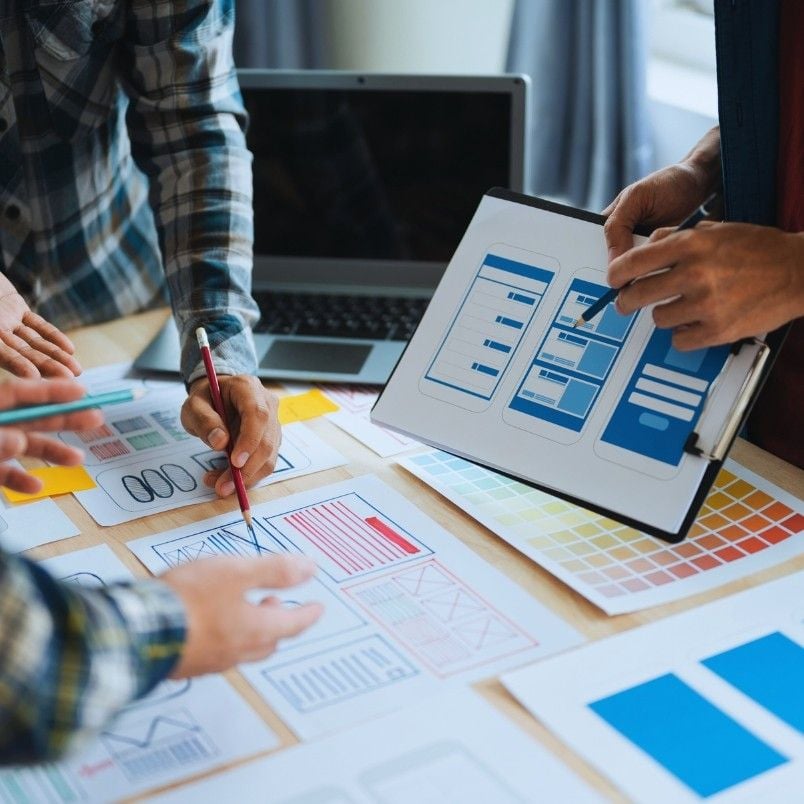 Three people collaborate around a desk with design sketches, a color palette, and a clipboard displaying a mobile app wireframe. One person points at the wireframe as others discuss Epic Examples and sketch ideas together.