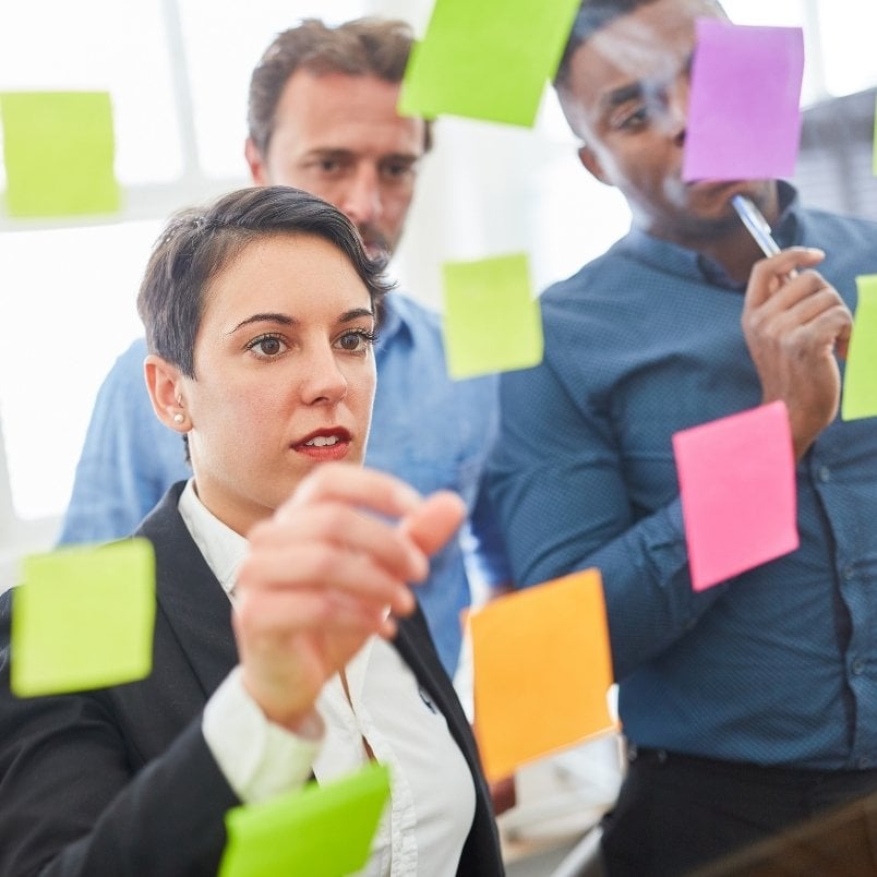 Three people stand behind a glass wall covered in colorful sticky notes, brainstorming and discussing ideas like "What Is An Epic In Scrum." One woman in front is placing a note while the two men behind her observe thoughtfully.