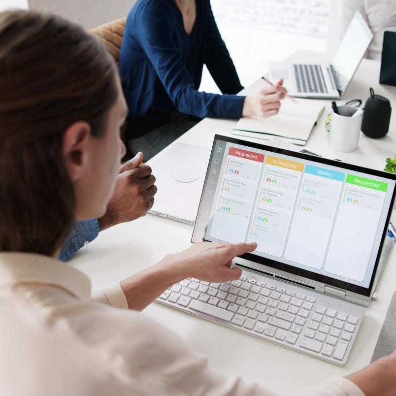 A person points at a Project Management Software for Small Businesses dashboard on a laptop screen during a team meeting, with two colleagues and another laptop visible on the table.