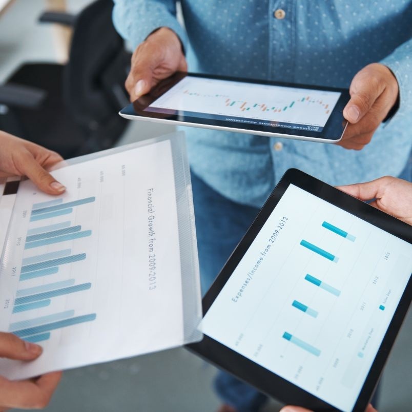 Three people hold tablets and a printed chart displaying bar graphs and data, collaborating as they review financial analytics and discuss Project Progress Tracking in BVDash in a professional setting.