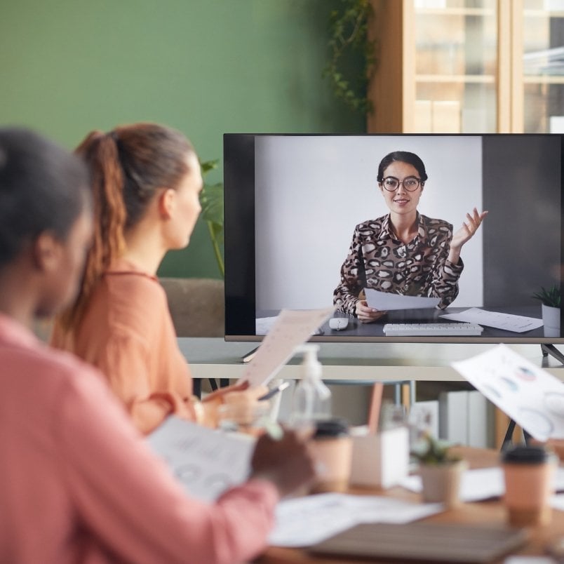 Two people sit at a table holding papers, discussing Project Management for Remote Teams while watching a woman speak on a video call displayed on a monitor in a modern office setting.