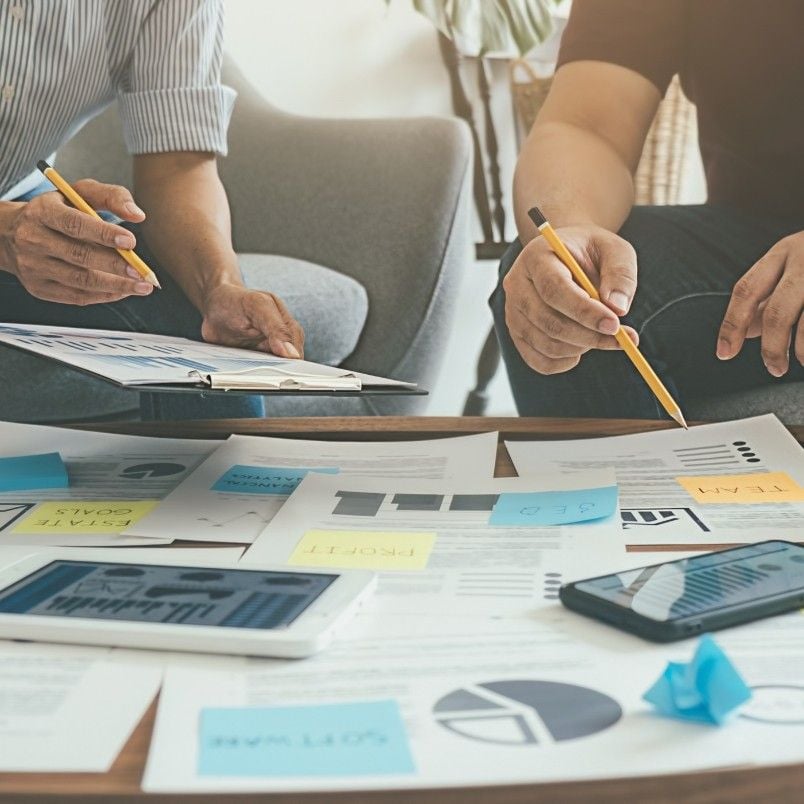 Two people sit at a table covered with charts, graphs, documents, a Stakeholder Map, and sticky notes, discussing and taking notes with pencils—suggesting a collaborative work or planning session.