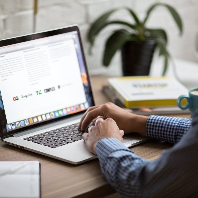 A person in a checkered shirt types on a laptop at a wooden desk with books, a mug, and a potted plant nearby. The laptop screen displays text and logos focused on Project Management for Remote Teams.