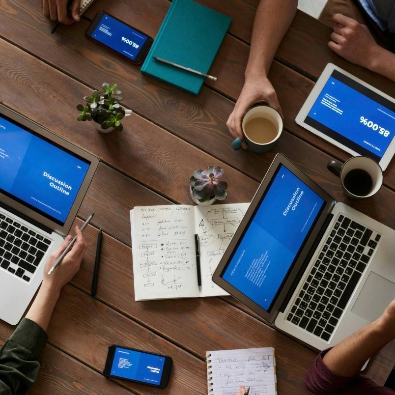 Four people sit around a wooden table with laptops, tablets, and smartphones displaying blue screens. Over coffee, they review notes on How to Write an Epic, preparing for a collaborative discussion outlined on their digital devices.