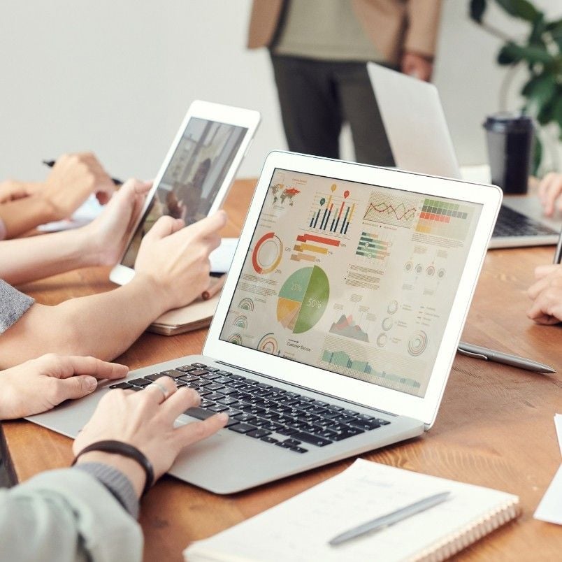 People sitting around a table using laptops and tablets, viewing colorful graphs and charts on a laptop screen; notebooks, papers, and a coffee cup are also on the table as they discuss Project Progress Tracking in BVDash.