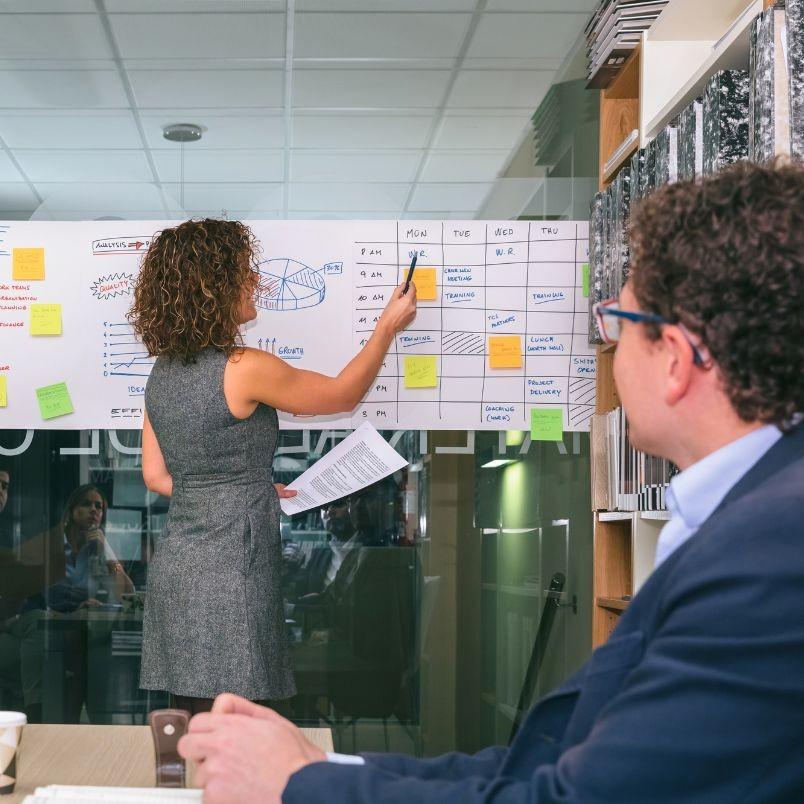 A woman stands in front of a whiteboard covered with charts, graphs, and sticky notes, pointing to a weekly schedule that highlights Project Progress Tracking in BVDash, while a seated man in glasses listens attentively in a modern office setting.