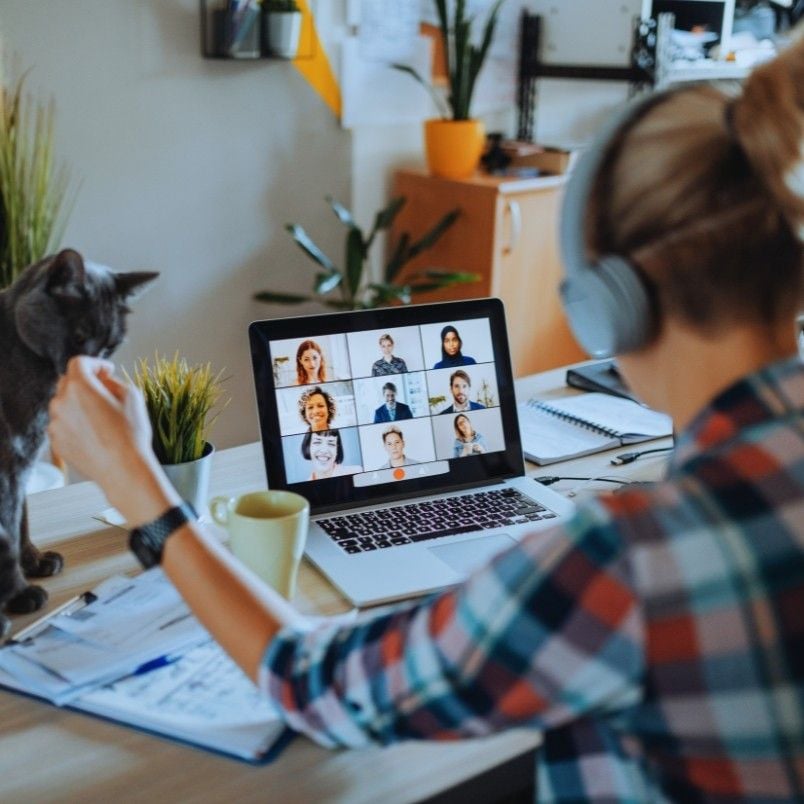 A woman wearing headphones sits at a desk with a laptop showing a virtual collaboration. She gestures with her hand while a cat sits nearby. The desk has a notebook, coffee mug, and a potted plant.