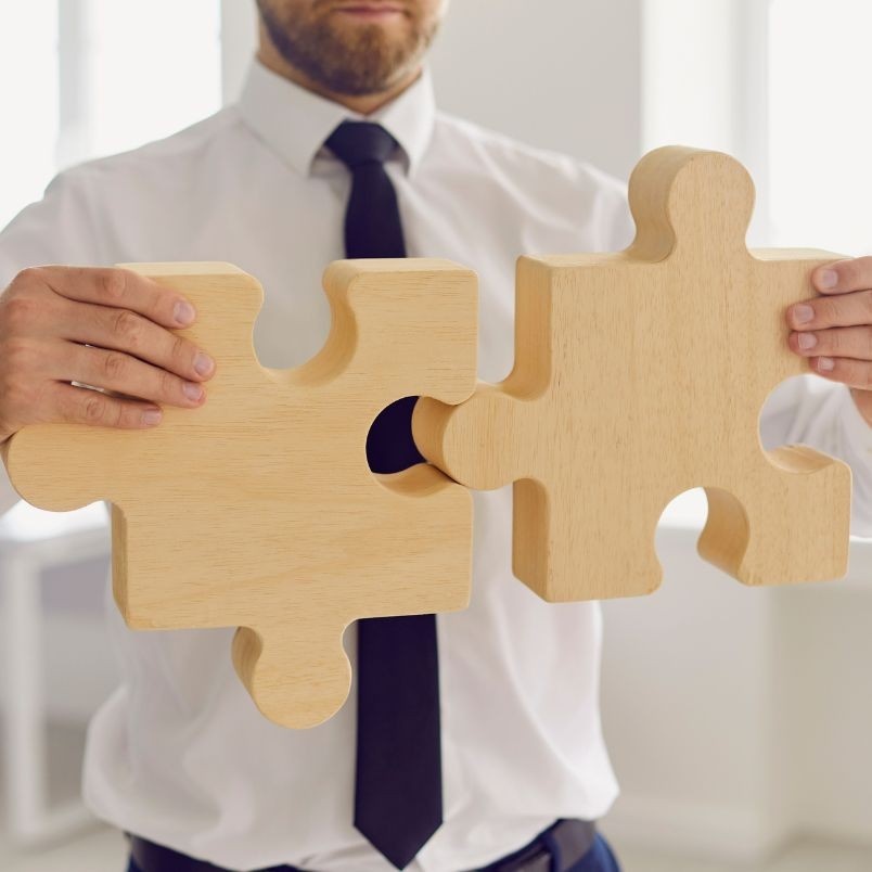 A man in a white shirt and black tie holds two large wooden puzzle pieces, fitting them together, symbolizing connection or problem-solving—much like Project Progress Tracking in BVDash brings solutions together.