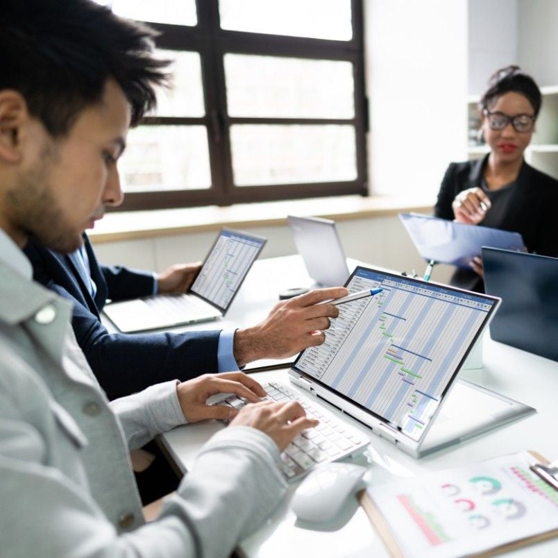 Three professionals collaborate at a table, utilizing Project Management Software for Small Businesses on their laptops—one types, another points at the screen displaying charts, while the third reviews documents in the background.