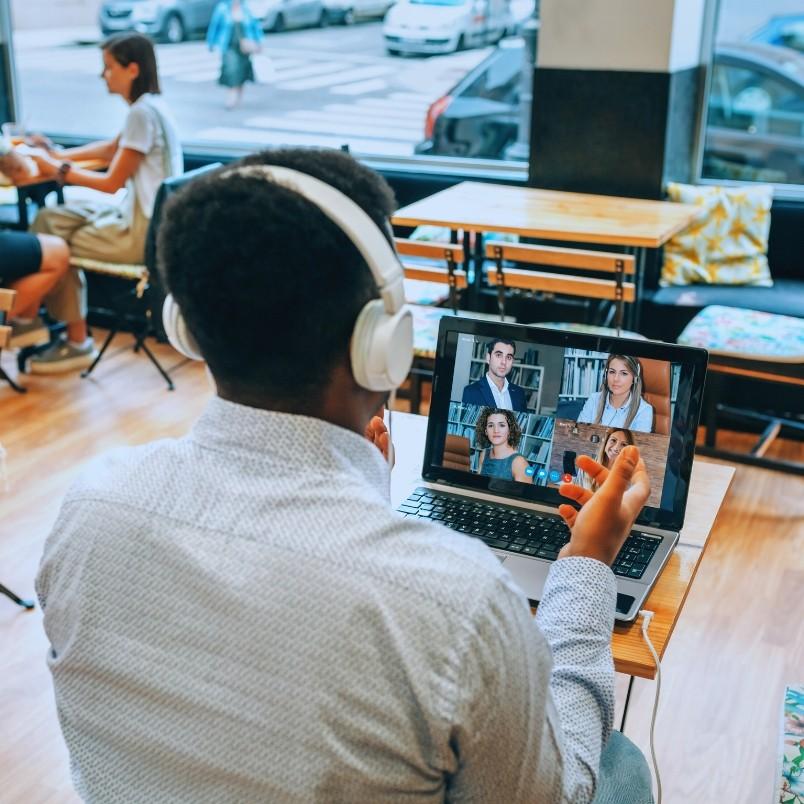 A person wearing headphones sits in a café, engaged in virtual collaboration on a video call with three others on their laptop, while other customers linger near large windows facing the street.