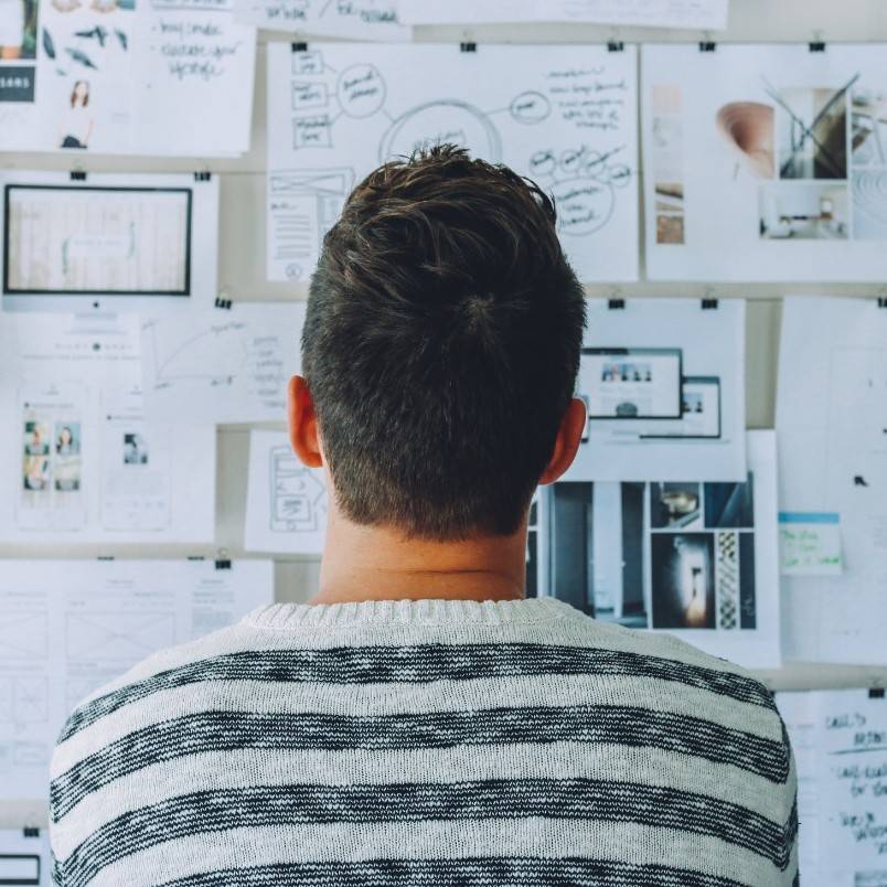 Person with short brown hair wearing a striped sweater is seen from behind, looking at a wall covered with papers and diagrams, planning how to write an epic through brainstorming ideas and organizing story elements.
