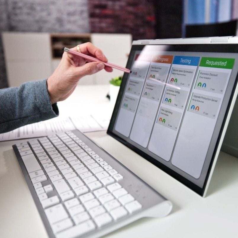 A person uses a stylus to interact with a touchscreen monitor displaying a digital task board from project management software for small businesses, with a wireless keyboard in the foreground on a modern office desk.